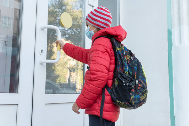 iskolás fiú orvosi maszkban közel van az ajtóhoz, és meghúzza a kilincset iskola előtt. Új valóság. Korlátozások a világjárvány megelőzésére. Egészségügyi koncepció - Fotó, kép