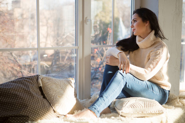 romantic brunette woman in sweater and jeans sitting on windowsill near window in apartment - Foto, immagini