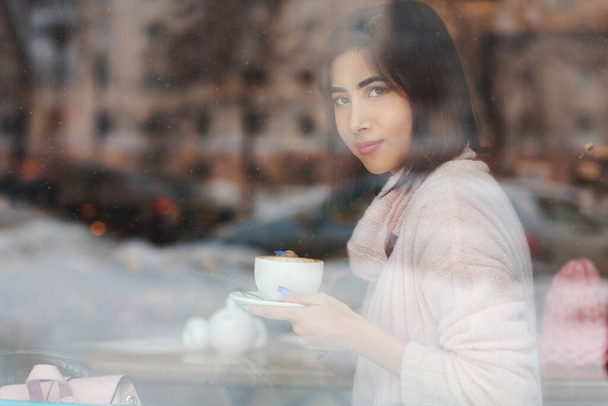 view trough window glass of beautiful brunette woman with coffee cup sitting at table in cafe - Zdjęcie, obraz