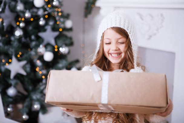 cute little girl in white warm winter clothes and hat with gift box posing in Christmas decorated room - Foto, imagen