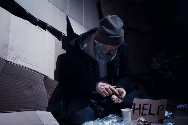 Бездомный бородатый сидит на коробках на улице и просит о помощи. Бездомный просит деньги на еду и ночлег. - Фото, изображение