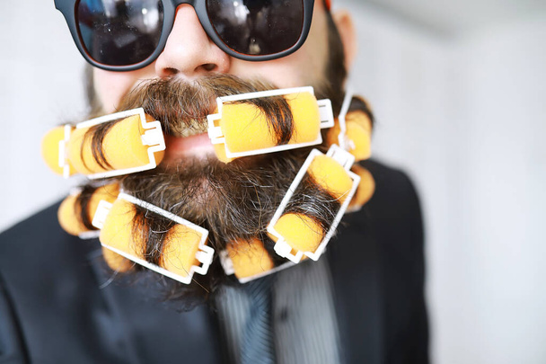 Fröhlicher Hipster-Mann mit Lockenwicklern im Bart. Überraschter Mann mit Brille blickt nach vorn. - Foto, Bild