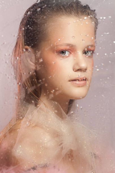 Young beautiful woman with perfect skin behind a glass with raindrops on pink background - Fotó, kép
