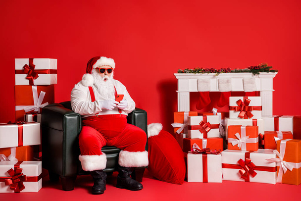 Portrait of his he nice attractive funky fat amazed Santa using device browsing web sitting in armchair isolated over bright vivid shine vibrant red color background - Фото, зображення