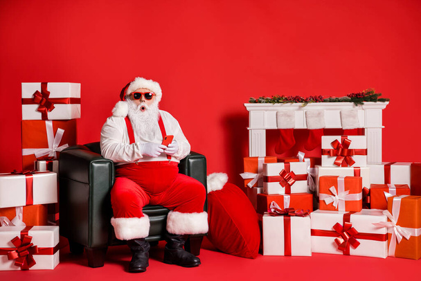 Portrét jeho on pěkný atraktivní funky tuk ohromený Santa otec pomocí přístroje app 5g sedí v křesle izolované přes světlé živé zářivá červená barva pozadí - Fotografie, Obrázek