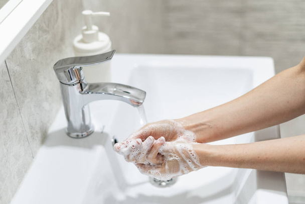 Covid és coronavirus pandémiás prevenció. Vágott kilátás nő mossa a kezét szappanhab a kerámia mosogató ellen víz áramlását, állt a fürdőszobában - Fotó, kép