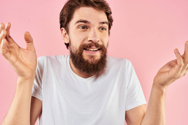 Бородатый мужчина в белой футболке жестикулирует с выражением лица рук вблизи розового фона - Фото, изображение