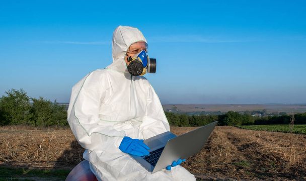 Fehér védőfelszerelést, kémiai maszkot és szemüveget viselő tudós laptopot használ a mezőgazdasági területen. - Fotó, kép
