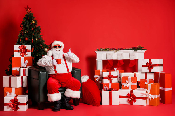 Portrait of his he nice attractive funky fat cheerful Santa sitting in armchair drinking beverage showing thumbup pause break isolated bright vivid shine vibrant red color background - Valokuva, kuva
