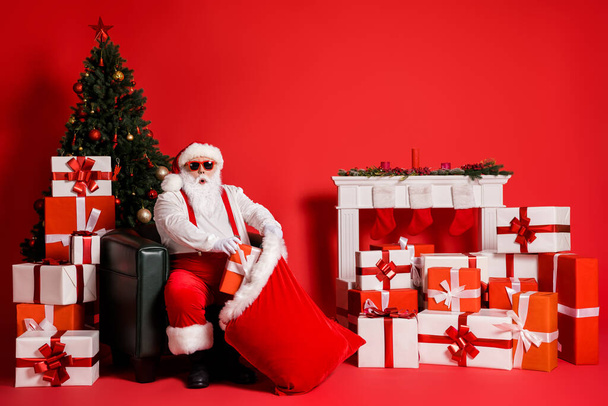 Portrait of his he attractive funky fat overweight amazed stunned Santa sitting in armchair decorated living-room packing giftboxes in sack isolated bright vivid shine vibrant red color background - Foto, Imagen