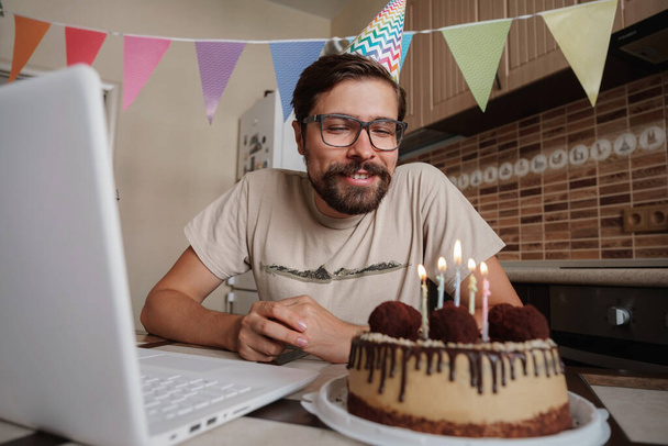 Férfi ünnepli születésnapját online karantén idő. A szülinapját ünnepli, elfújja a gyertyát a szülinapi tortán, és videóhívást kezdeményez. Coronavirus járvány 2020. - Fotó, kép