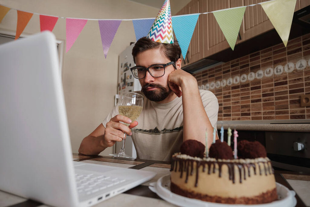 Rozrušený mladý muž sedící u narozeninového dortu a dívající se na něj smutnýma očima. Koncept samoty v karanténě během koronavirové pandemie COVID-19. - Fotografie, Obrázek