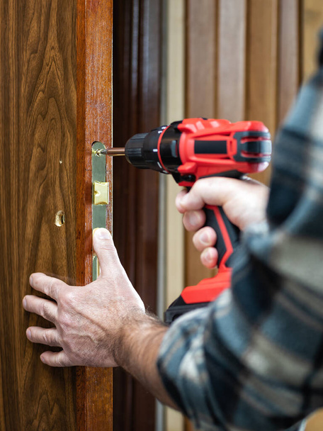 Primer plano. Carpintero con un destornillador eléctrico fija la cerradura de una puerta de madera. Industria de construcción.  - Foto, imagen