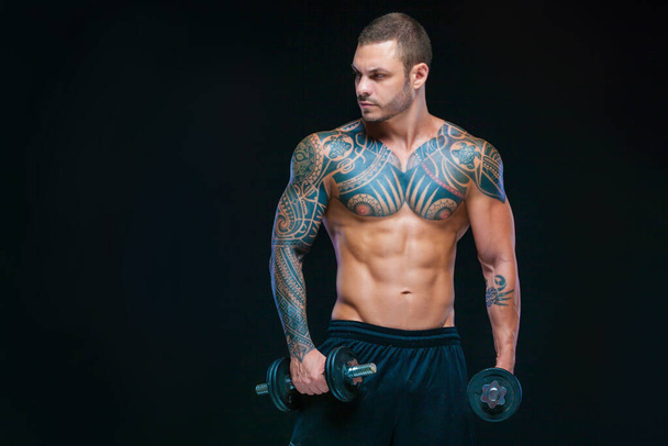 Tattooed strong muscular athletic man pumping up muscles with dumbbells on black background - Zdjęcie, obraz