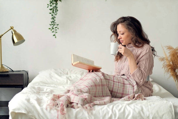 Young woman with cup of hot beverage reading book in bed - Foto, afbeelding