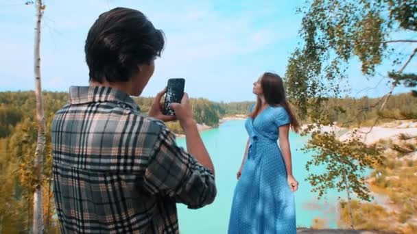 Egy férfi, aki a barátnőjét fotózza a telefonján a kék tó és erdő hátterében - életmód - Felvétel, videó