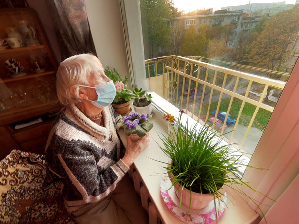 The second wave. Covid-19, health, safety and pandemic concept - senior old lonely woman wearing protective medical mask sitting near the window at home for protection from virus - Photo, Image