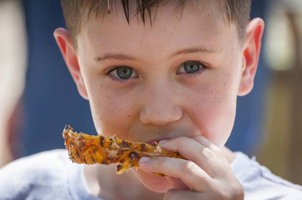 Cute Caucasian 8 year old boy with blue eyes eating pineapple looking at the camera close up portrait image. - Photo, Image
