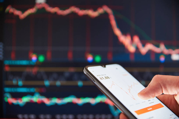 Persoon controleert de aandelenkoersen op een smartphone voor een groot scherm markttarieven. Persoon die aandelenkoersen bekijkt op een smartphone met marktprijzen op een scherm op de achtergrond. - Foto, afbeelding