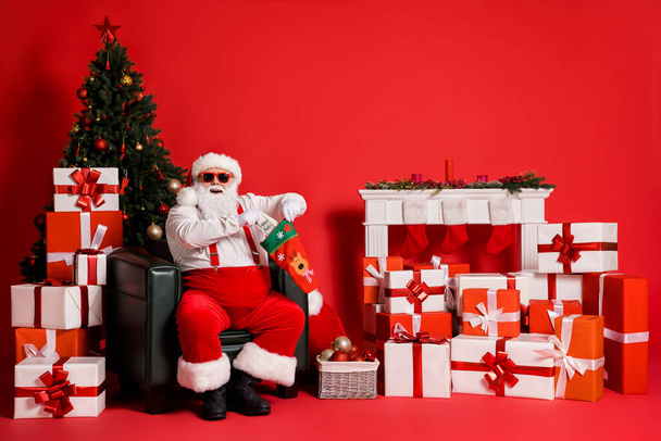Portrait of his he attractive fat overweight cheerful glad Santa sitting in armchair living-room packing us cash gift in ugly sock isolated bright vivid shine vibrant red color background - Foto, Imagem