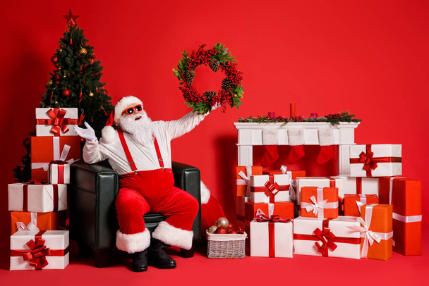 Nice attractive fat overweight cheerful cheery glad Santa sitting in armchair holding in hands festal wreath decor having fun isolated bright vivid shine vibrant red color background - Фото, зображення