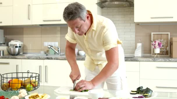 Çekici gri saçlı adam evde yemek pişiriyor, mutfak masasında salata için sebze kesiyor. - Video, Çekim