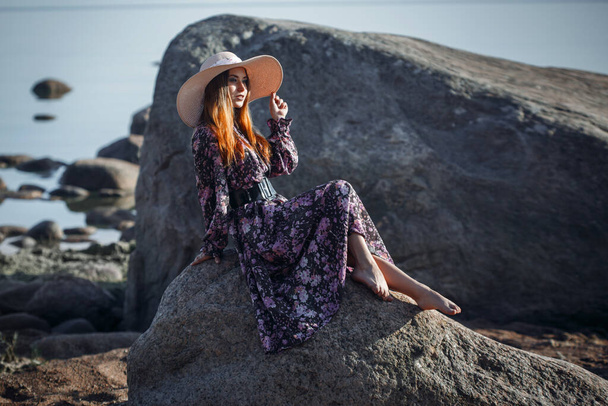 Beautiful bright girl in a big straw hat is resting sitting on a stone on a wild sea shore. - Zdjęcie, obraz