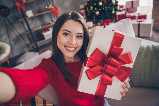 Φωτογραφία του θετικού κορίτσι κρατήσει κουτί δώρου κάνουν selfie κάθονται καναπέ στο σπίτι σε εσωτερικούς χώρους με χριστουγεννιάτικη διακόσμηση x-mas - Φωτογραφία, εικόνα