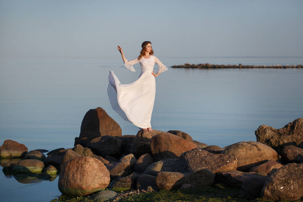 Hermosa novia chica tierna en un vestido blanco largo se encuentra en las piedras en la orilla del mar por la noche. - Foto, Imagen