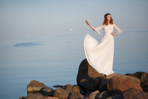 Beautiful tender girl bride in a long white dress stands on the stones on the seashore in the evening. - Zdjęcie, obraz