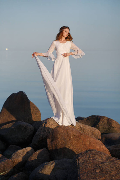 Beautiful tender girl bride in a long white dress stands on the stones on the seashore in the evening. - Foto, immagini