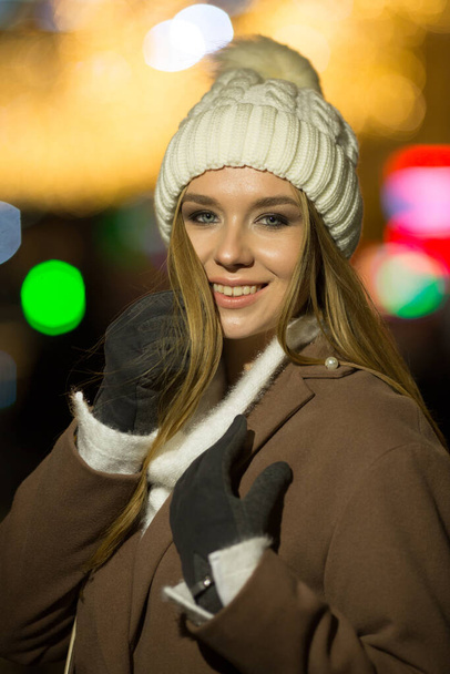 beautiful girl, in the evening, against the background of lights, a white hat and a beige coat. smiling at camera, close-up portrait - Foto, Imagen