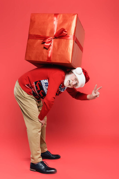 Senior man in santa hat showing peace sign while holding gift on back on red background - Фото, изображение