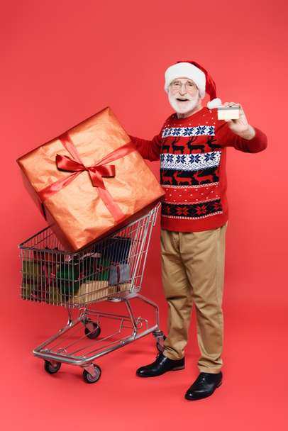 Cheerful senior man in santa hat showing credit card near shopping cart with presents on red background - Foto, imagen
