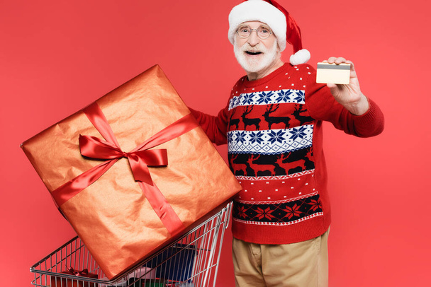 Smiling elderly man in santa hat holding credit card near gifts in shopping cart on red background - Foto, immagini