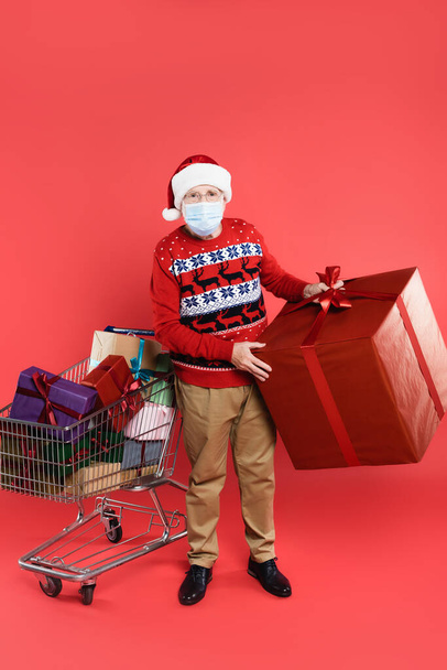 Senior muž v lékařské masce a Santa klobouk držení dárkové krabice v blízkosti nákupního košíku s dárky na červeném pozadí - Fotografie, Obrázek