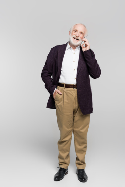 Smiling elderly man with hand in pocket of pants talking on smartphone on grey background - Foto, afbeelding