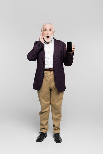 Shocked elderly man with hand near head holding smartphone with blank screen on grey background  - Foto, Imagem