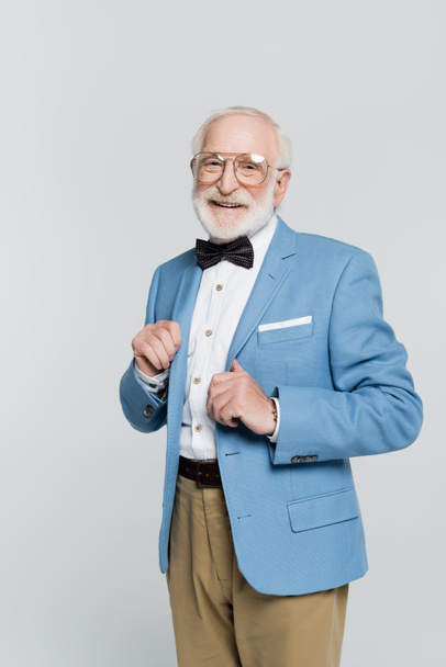 Smiling elderly man in eyeglasses and bow tie looking at camera isolated on grey  - Foto, imagen