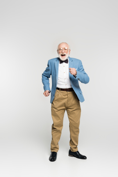 Cheerful senior man in jacket and bow tie dancing on grey background  - Фото, зображення