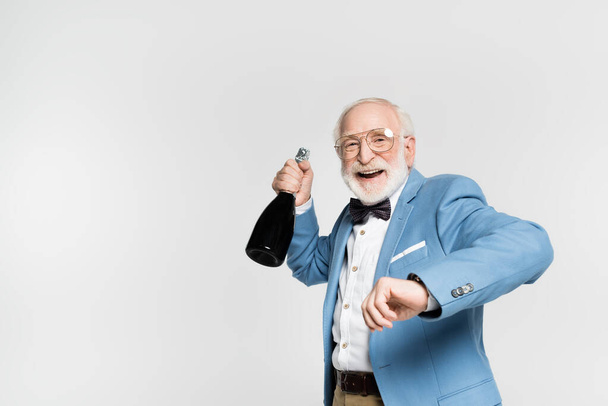 Positive elderly man in jacket and bow tie holding bottle of champagne isolated on grey  - Foto, afbeelding