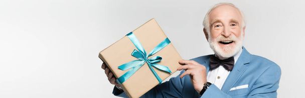 Uomo anziano in papillon e giacca contenente scatola regalo e sorridente alla fotocamera isolata su grigio, banner - Foto, immagini