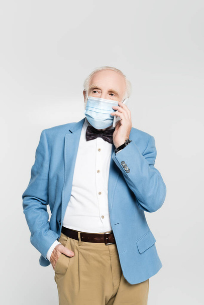 Starší muž v lékařské masce a bundě mluví na smartphone izolované na šedé  - Fotografie, Obrázek