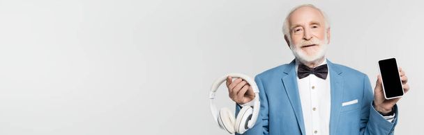 Χαμογελώντας ηλικιωμένος με παπιγιόν και σακάκι κρατώντας ακουστικά και smartphone με λευκή οθόνη απομονώνονται σε γκρι, πανό  - Φωτογραφία, εικόνα