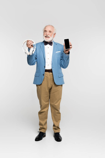 Smiling elderly man holding wireless headphones and smartphone on grey background  - 写真・画像