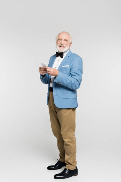 Smiling elderly man in bow tie and jacket holding smartphone on grey background  - Foto, afbeelding