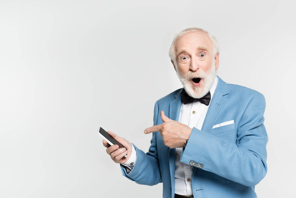 Emocionado hombre mayor en pajarita y chaqueta apuntando con el dedo al teléfono inteligente aislado en gris  - Foto, imagen