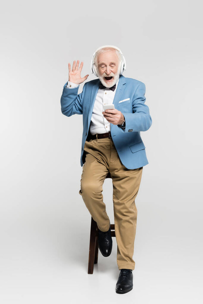 Excited senior man in headphones waving hand at smartphone on chair on grey background  - Zdjęcie, obraz