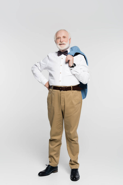 Smiling senior man with hand on hip holding jacket on grey background  - Foto, afbeelding