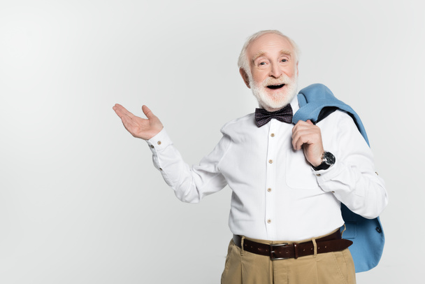 Hombre anciano sonriente con corbata de lazo sosteniendo chaqueta y señalando con la mano aislada en gris  - Foto, Imagen